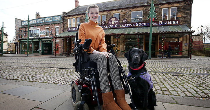 disabled blogger kate stanforth sat in wheelchair with assistance dog next to her in Beamish Museum 1900s town street.